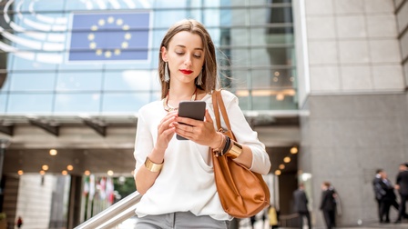 Person mit Schmuck behangen blickt auf Smartphone, im Hintergrund verschwommen weitere Personen und Gebäude mit EU-Zeichen, gelbe Sterne im Kreis verlaufend auf dunkelblauem Hintergrund