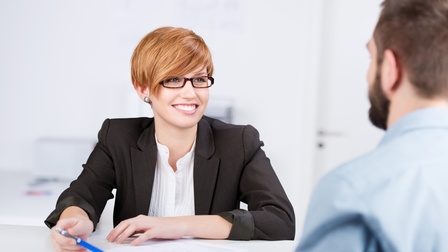 Frontale Aufnahme einer Person in einem Sakko, mit Brille und Bluse, die an einem Tisch sitzt und sich mit einer Person unterhält, die ihr gegenüber sitzt. Vor ihr liegt ein Blatt Papier