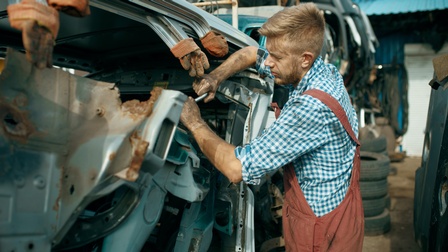 Person in Arbeitslatzhose schraubt an verschrottetem Fahrzeug