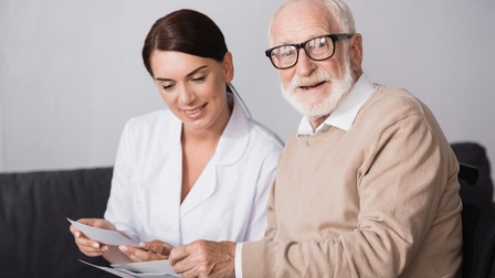 Pensionierte Person mit Brille und weißen Haaren und Bart betrachtet freudig gemeinsam mit einer Person in weißer Dienstkleidung Bilder vergangener Tage