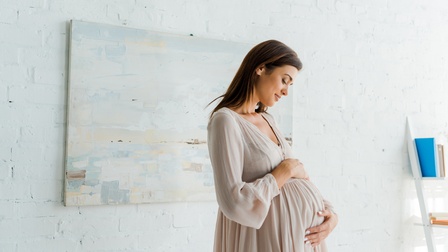 Eine Person mit offenen, dunklen Haaren in einem hellen Kleid steht vor einer hellen Wand. Die Person hat ihre Hände auf ihrem runden Buch und blickt darauf