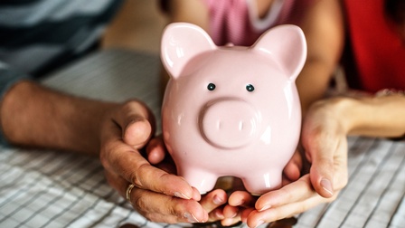 Mehrere Hände halten ein Rosa Sparschwein, darunter liegen Geldmünzen auf einem Tisch
