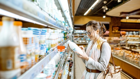 Seitliche Aufnahme einer Person mit Brille, die neben einem Kühlregal in einem Supermarkt steht und von einer weißen Plastikflasche abliest. Im Hintergrund sind weitere Regale und eine Theke