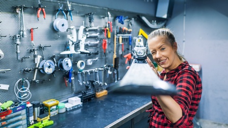 Person in kariertem Hemd hält Schi mit beiden Händen vor sich, kneift eine Auge zu und lugt mit anderem entlang des Schis, im Hintergrund Werkzeugwand mit Werkbank