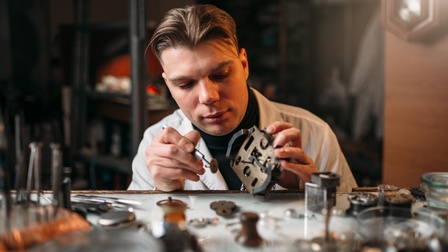 Person sitzt an Werktisch und setzt Kleinteil in Uhr ein, ringsum Pinzetten, Werkzeuge und Bestandteile von _Uhren