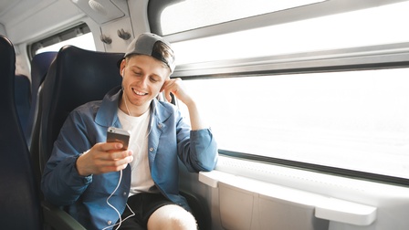 Portrait einer lächelnde Person mit umgekehrt aufgesetzter Kappe und Kopfhörern auf Smartphone in Hand blickend sitzt auf Platz in Zug neben Fenster und stützt an kleinem Tischchen Arm ab, auf den Kopf gestützt ist