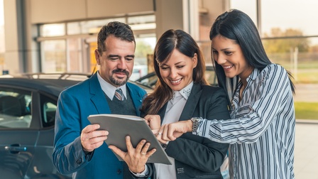 Drei lächelnde Personen blicken auf Katalog, im Hintergrund Autos