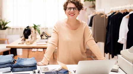 Lächelnde Person mit Brillen steht an Tisch mit aufgeklapptem Laptop, Zahlungsgerät und Stapeln von Jeans, im Hintergrund Kleiderstangen und Tisch mit modischen Accessoires