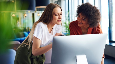 Zwei Personen stehen vor einem Bildschirm und beugen sich mit ihren Oberkörpern leicht nach vorne. Die linke Person deutet mit ihrem Stift auf den Bildschirm und blickt darauf. Die rechte Person blickt die linke Person an