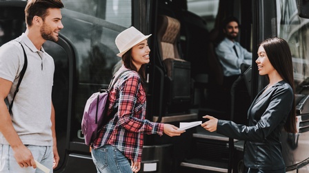 Zwei Personen stehen sich vor einer geöffneten Bustür gegenüber. Die linke Person übergibt der rechten Person einen Zettel. Hinter der linken Person steht eine weitere Person. Im Bus sitzt eine Person direkt hinter der Glasscheibe.