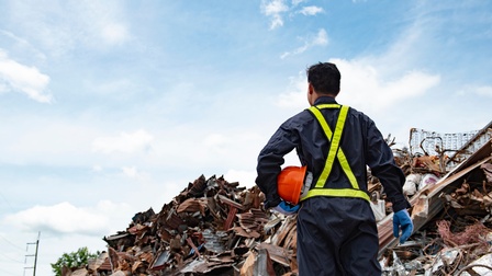 Rückenansicht einer Person in einem dunkelblauem Overall, mit Handschuhen und Bauhelm in der linken Hand, die gegenüber eines Müllhaufens steht. Dahinter ist der bewölkte Himmel