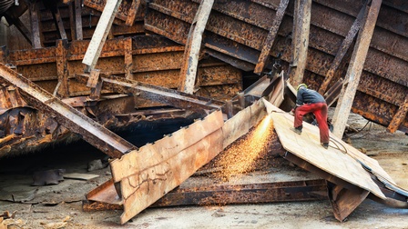 Person mit Schutzhelm in Rückenansicht auf großem Blech stehend Schweißgerät bedienend, ringsum große Stücke rostigen Metalls von einem Schiff