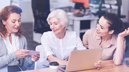 Personen unterschiedlicher Generationen sitzen bei einem Tisch zusammen und unterhalten sich, eine ältere Person mit weißen Haaren arbeitet dabei mit einem Laptop, im Hintergrund zeigt sich eine Büroräumlichkeit