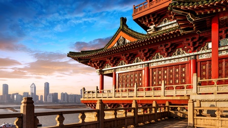 Rechts im Vordergrund ist ein historisches Gebäude mit roten Säulen und spitzen Dächern. Daneben ist ein Gewässer. Am Horizont ist die Skyline der Stadt Peking mit verschiedenen Häusern. Am Himmel sind ein paar Wolken