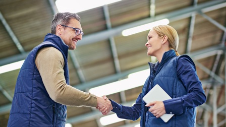 Personen in blauer Arbeitskleidung lächeln sich an und geben sich die Hand in einer Betriebshalle