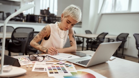 Eine Person arbeitet in einem Büro an grafischen Designs. Am Tisch liegen Zettel mit unterschiedlichen Farben und ein aufgeklappter Laptop.