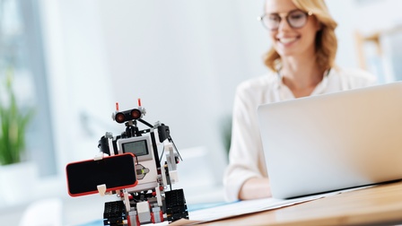 Lächelnde Person mit Brille sitzt mit Laptop vor einem selbst automatisiertem Roboter bei einem Tisch, der ein Smartphone transportiert