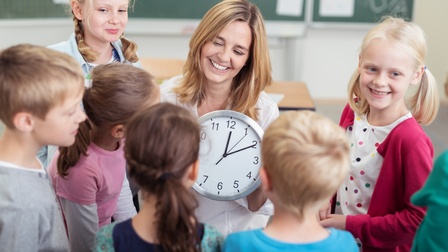 Person mittleren Alters hält eine Uhr in Händen und Kinder stehen ringsherum, im Hintergrund stehen Tische und eine grüne Tafel