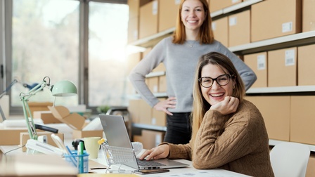 Zwei E-Commerce-Geschäftspersonen arbeiten im Büro. Auf dem Schreibtisch befinden sich ein Laptop, Kartons und Büromaterialien. Im Hintergrund sind Regale voller Kartons zu erkennen.