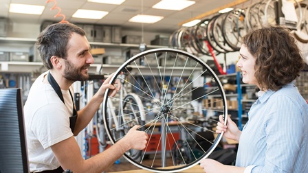 Zwei Personen in Fahrradgeschäft halten jeweils an einer Fahrradspeiche die auf Tresen aufsetzt und lächeln sich an