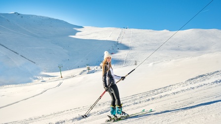 Seitliche Aufnahme einer Person in Skikleidung, Skischuhen und Ski, die sich an einer schrägen Stange festhält, die an einem Seil befestigt ist und direkt in die Kamera blickt. Unter ihr und hinter ihr ist Schnee