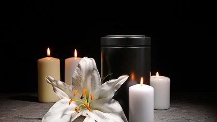 Schwarze Urne umrandet von vier weißen brennenden Kerzen und Blüte einer weißen Lilie
