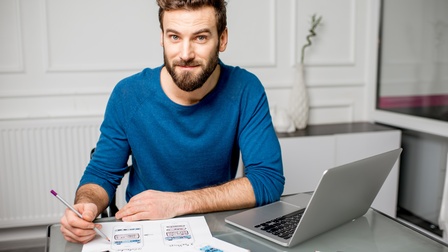 Person mit kurzen braunen Haaren,  Bart und blauem Pullover sitzt bei einem Schreibtisch mit Laptop und zeichnet mit Stiften ein UX Design auf Zetteln