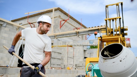 Person mit Schutzkleidung arbeitet bei Mischmaschine am Bau und fertigt einen Ziegelbau