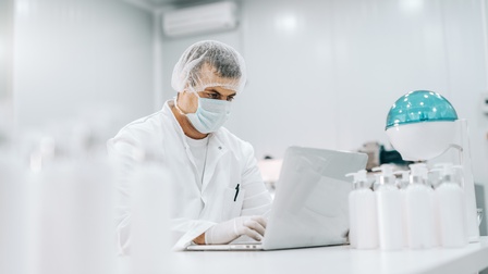 Person mit Mundnasenschutz, Haarnetz und Schutzhandschuhen tippt auf Notebook in Labor