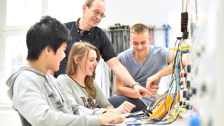 Seitliche Aufnahme von drei jungen Personen, die an einem Tisch sind. Vor ihnen sind verschiedene Kabel. Neben ihnen steht eine Person, die auf die Kabel deutet