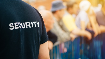 Rücken einer Person im Ausschnitt im Fokus mit schwarzem T-Shirt mit der Aufschrift Security, im Hintergrund verschwommen Personen an Zaun lehnend