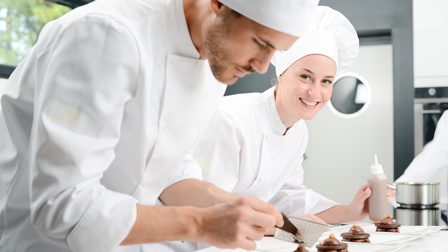 Zwei Personen in weißen Kochgewändern mit Kochhauben platzieren und dekorieren Süßspeisen auf Tellern