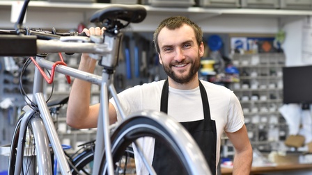 Lächelnde Person in Arbeitsschürze steht in Fahrradwerkstatt und hält Hand auf silbernes Fahrrad