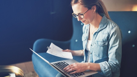Person mit Brillen sitzt auf blauem Sofa und hält in einer Hand Dokumente, mit der anderen Hand tippt sie auf Laptoptastatur