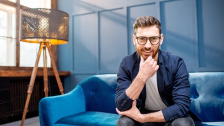 Lächelnde Person mit dunklem Bart und Brillen sitzt auf blauer Couch und hält eine Hand ans Kinn, im Hintergrund Fenster, Wandvertäfelung und eine Stehlampe