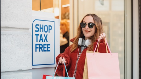 Eine Person mit Sonnenbrille, Kopfhörer und vielen Einkaufstaschen in beiden Händen steht vor dem Eingang eines Geschäfts. Neben ihr an der Wand ist ein Schild montiert, auf dem Shop Tax Free steht