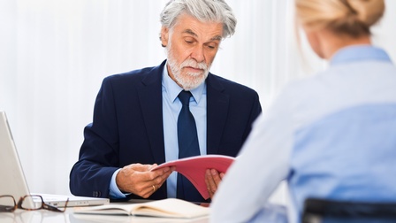 Eine Person schaut die Bewerbungsmappe einer zum Bewerbungsgespräch eingeladenen Person durch, die gegenüber von ihr sitzt.