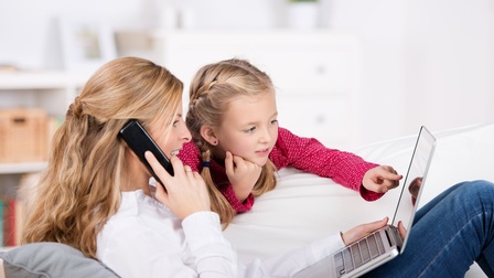Eine Erwachsene telefoniert auf der Couch liegend am Smartphone und hat einen Laptop aufgeklappt, daneben deutet ein Kind mit dem finger auf den Bildschirm