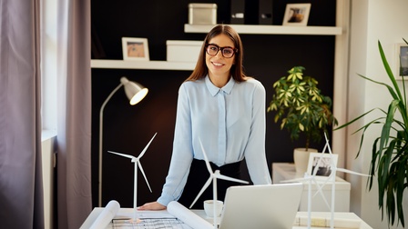 Halbporträt einer Person mit heller Bluse und Brille, die sich leicht nach vorne beugt und am vor ihr stehenden Tisch abstützt. Auf dem Tisch sind eine teilweise ausgerollte Papierrolle, ein aufgeklappter Laptop und kleine Windräder