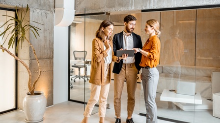 Frontale Aufnahme dreier Personen in Bürokleidung, die nebeneinander vor einer Glaswand in einem Bürogebäude stehen. Die linke Person hält ein Tablet, auf dessen Bildschirm die anderen beiden Personen deuten