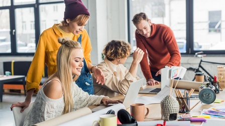 Zwei Personen sitzen an einem Tisch. Zwei Personen stehen jeweils neben einer sitzenden Person. Die Personen interagieren miteinander. Auf dem Tisch sind Stifte, Papierrollen, Unterlagen sowie zwei aufgeklappte Laptops