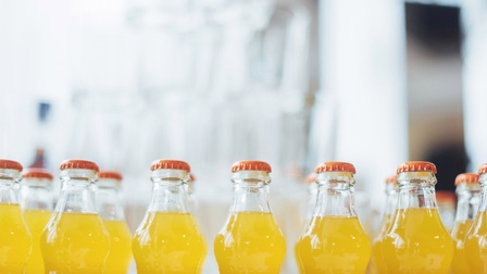 Mehrere transparente Glasflaschen mit orangen Kronkorken und gelber Flüssigkeit nebeneinander stehend, Hintergrund verschwommen