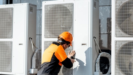 Eine Person in einer orange-schwarzen Jacke, orangem Helm und Sonnenbrille steht neben einem Außengerät einer Klimaanlage und hält einen Schraubenzieher darauf