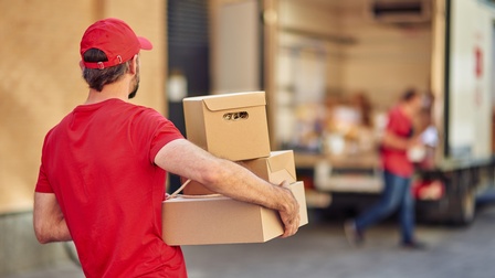 Rückenansicht einer Person in roter Kappe und rotem Oberteil, die drei Kartons mit beiden Händen hält. Im Hintergrund steht ein Lastwagen mit geöffneter Tür hinten