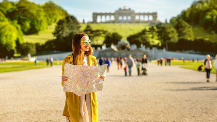 Eine Person mit einem Stadtplan und Sonnenbrille steht auf einem sehr breiten Kiesweg. Im Hintergrund sind verschiedene Leute. Im Hintergrund ist ein grüner Hügel, auf dem ein historisches Gebäude steht