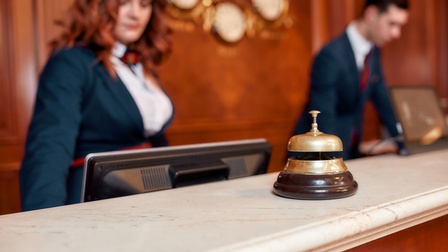 Im Fokus ist eine Hotelglocke, die auf einem Tresen steht. Hinter dem Tresen stehen zwei Personen in Anzügen. Die linke Person blickt auf einen Bildschirm. Hinter ihnen ist eine vertäfelte Wand