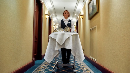 Eine Person in einer hellen Bluse, einer Weste und einem Ausweisschild auf der Weste schiebt in einem Gang einen kleinen Wagen entlang. Auf dem Wagen stehen Teller, eine Kanne und eine Tasse