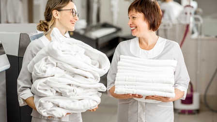 Zwei einander anlächelnde Personen in weißer Arbeitskleidung, eine Person hält Stapel weißer Morgenmäntel in Händen, die anderer Person trägt Stapel weißer Handtücher