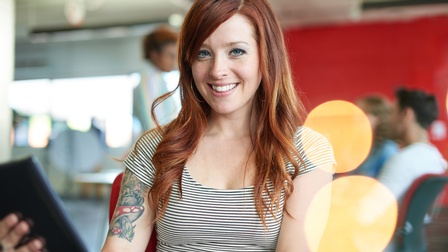 Person mit rötlichen langen Haaren trägt mit gestreiftem Shirt, Tattoo am rechten Oberarm blickt freudig in die Kamera und hält ein Tablet in der Hand, im Hintergrund  sitzen und stehen weitere Personen in einem roten Raum