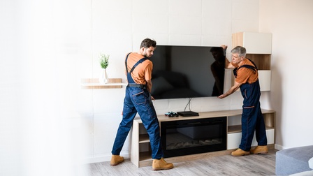 Zwei Personen mit kurzen Haaren und Bart sowie orangem Shirt und blauer Latzhose und beigen Schuhen halten einen Fernseher bei einer Wohnzimmerverkleidung